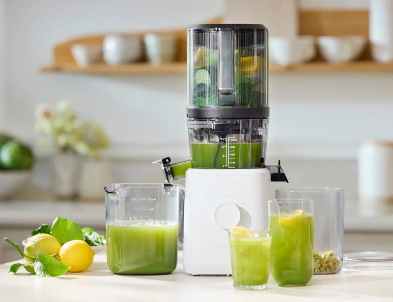 A white juicer filled with green fruits and vegetables sits on a kitchen counter, with two glasses and a pitcher of fresh green juice beside it. Lemons and leafy greens are also on the counter.