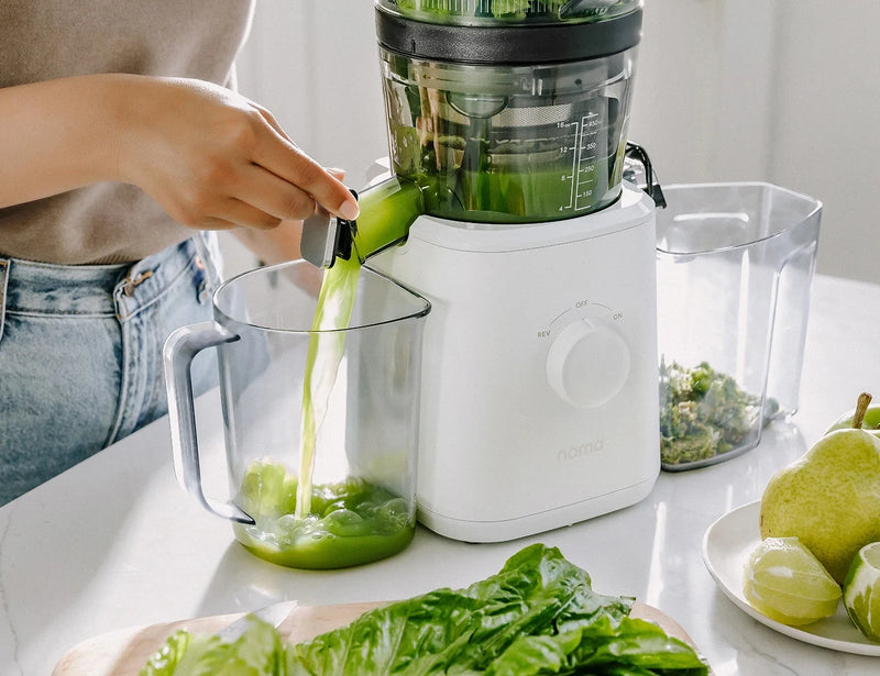 A person uses a white juicer to pour fresh green juice into a clear measuring pitcher. Green produce, including lettuce and pears, are on the counter nearby.