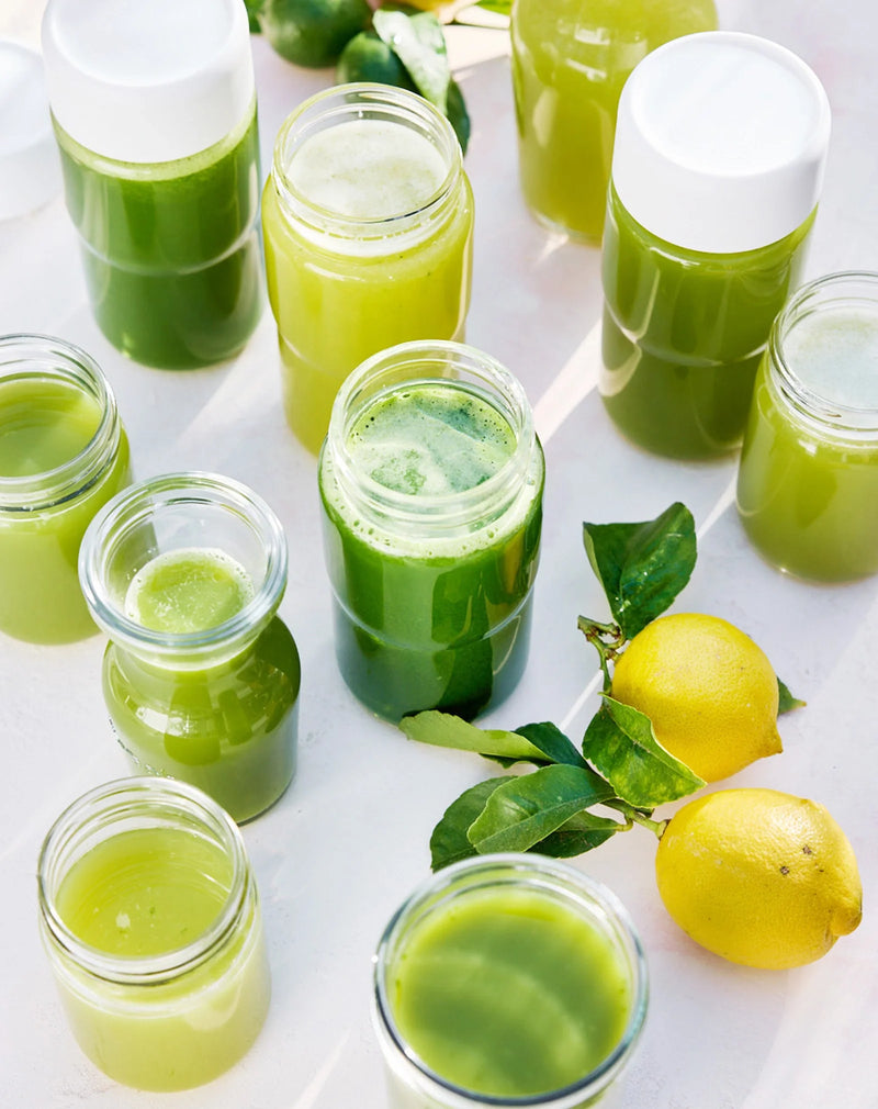 Assorted glass jars and bottles filled with green juice are arranged on a white surface, next to two fresh lemons with green leaves attached.
