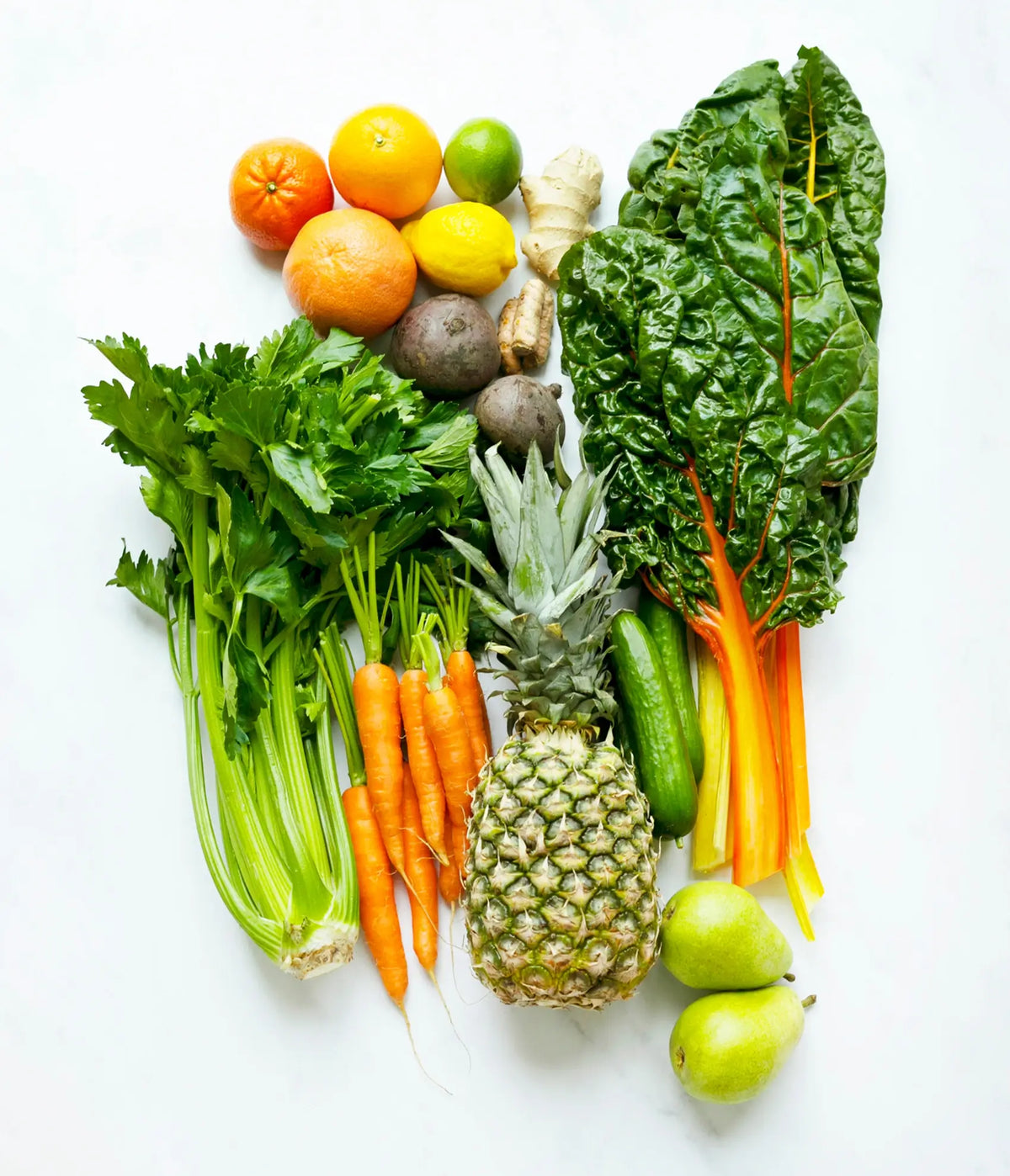 A colorful assortment of fresh fruits and vegetables, including carrots, celery, pineapple, pears, citrus fruits, cucumbers, beets, ginger, and rainbow chard, arranged on a white surface.