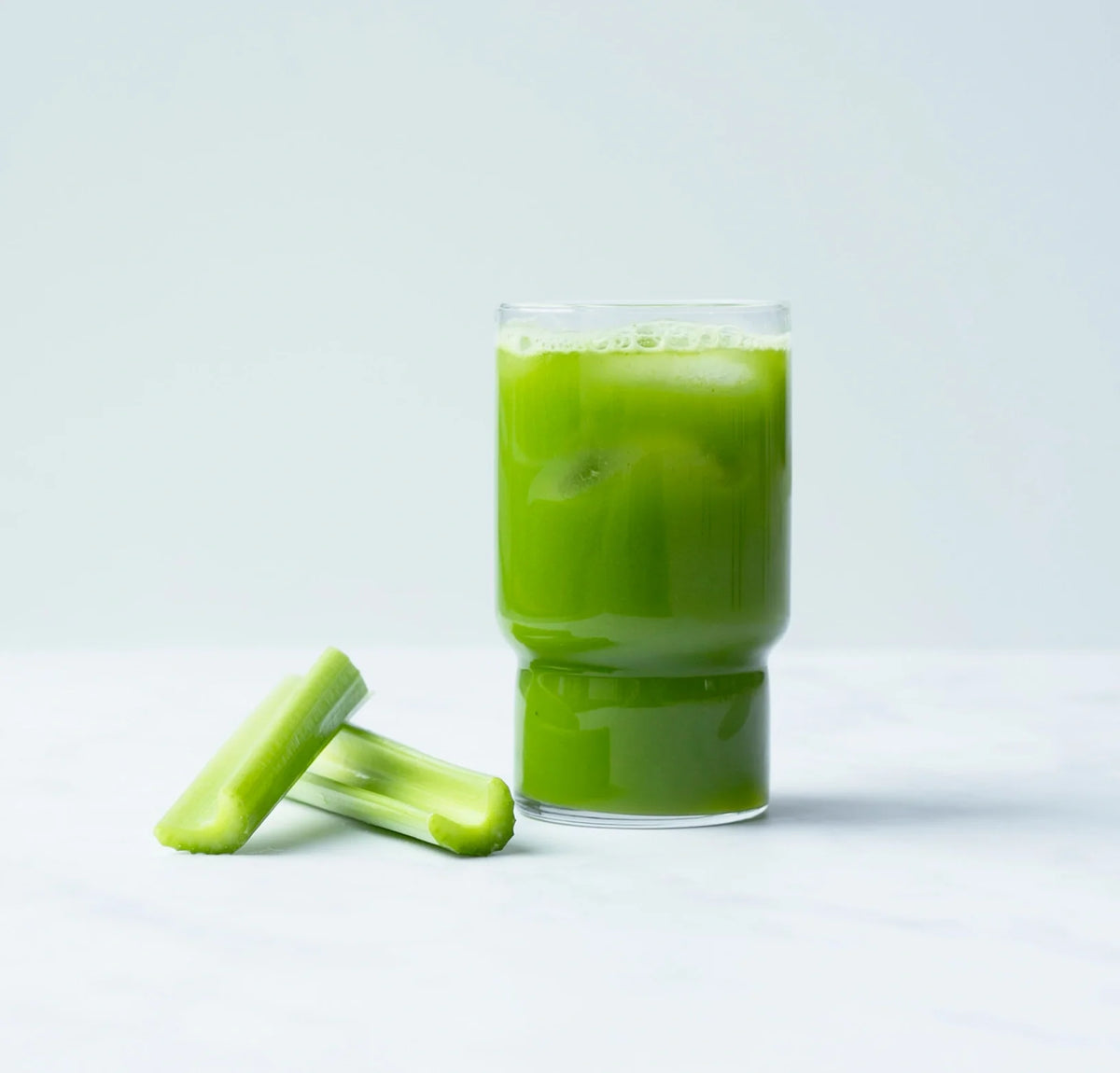 A glass of green juice sits on a white surface next to two pieces of fresh celery. The background is light and plain, highlighting the vibrant green color of the juice.