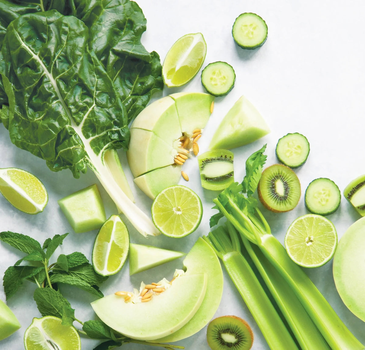 Sliced honeydew melon, celery, lime, cucumber, kiwi, Swiss chard, and mint leaves are arranged on a white surface. The fruits and vegetables are mostly green and displayed in a fresh, vibrant composition.