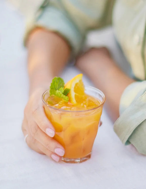 A person with light nail polish holds a glass of orange juice with ice, garnished with mint and an orange slice, on a white surface.