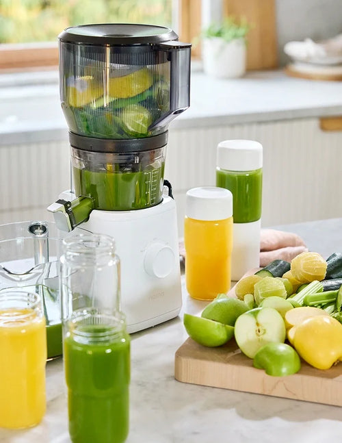A juicer on a kitchen counter processes limes, with bottles of green and yellow juice nearby. Fresh apples, lemons, cucumbers, and limes are on a wooden cutting board.