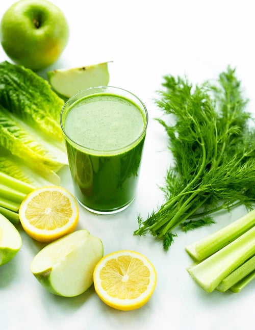 A glass of green juice surrounded by green apple slices, lettuce, dill, celery sticks, and halved lemons on a white surface.