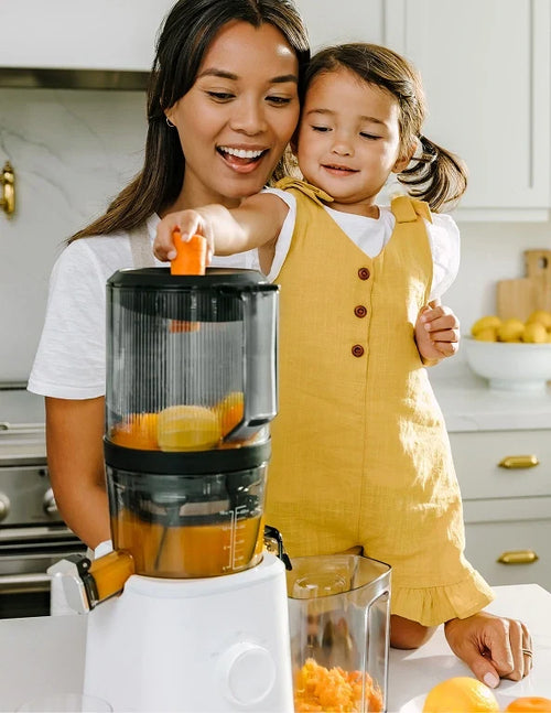A woman and a young girl in a yellow dress use a juicer in a bright kitchen. The girl helps by placing an orange piece into the juicer, while the woman smiles and watches. Fresh juice and fruit are visible nearby.