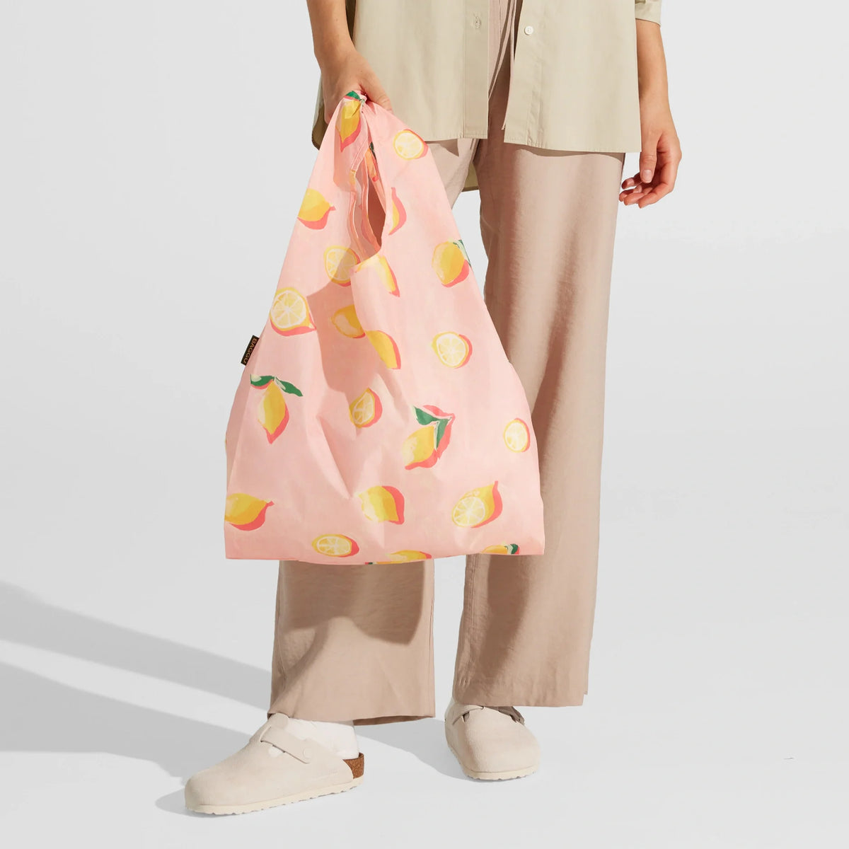 A person dressed in beige pants and a light shirt holds the Nama + Baggu Standard Tote Bag—a pink reusable grocery bag featuring a lemon and lemon slice design. They are also wearing light-colored slip-on shoes.