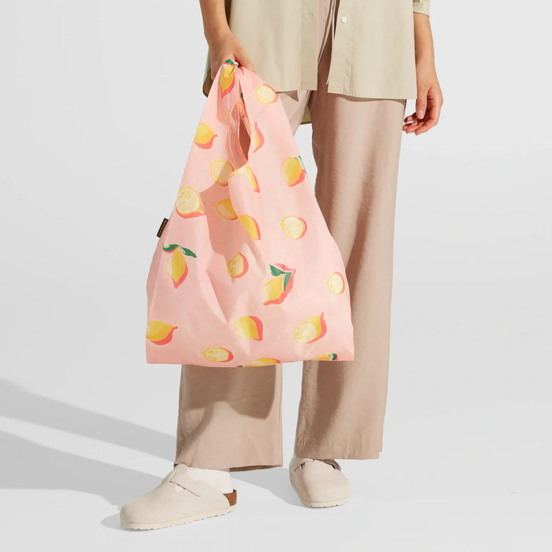 A person dressed in beige pants and a light shirt holds the Nama + Baggu Standard Tote Bag—a pink reusable grocery bag featuring a lemon and lemon slice design. They are also wearing light-colored slip-on shoes.