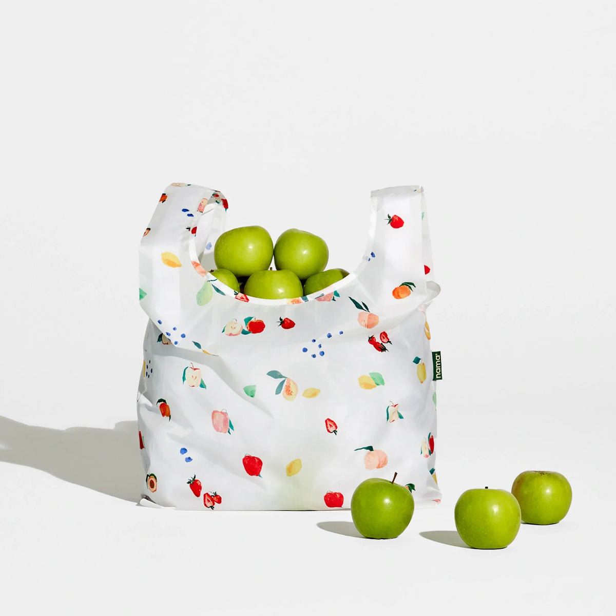 The Nama + Baggu Standard Tote Bag, featuring a white design with colorful fruit prints, holds several green apples to showcase fresh produce. Three additional green apples are arranged in front of the bag on a white background.