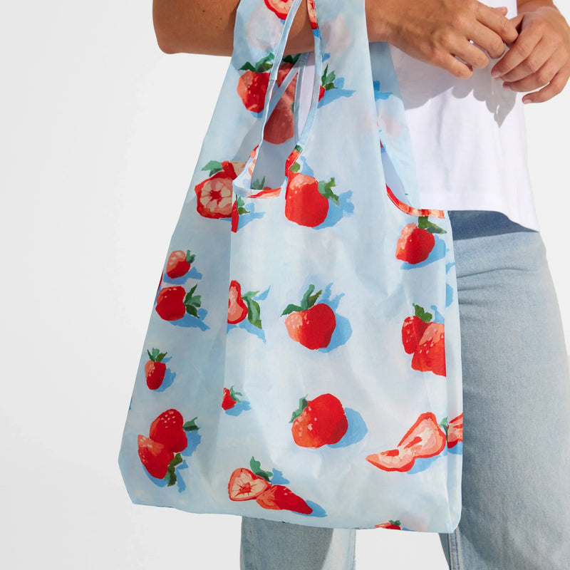 A person holds the Nama + Baggu Standard Tote Bag, a light blue reusable bag adorned with red strawberries and strawberry slices—perfect for carrying fresh produce. The person is dressed in a white shirt and light blue jeans.