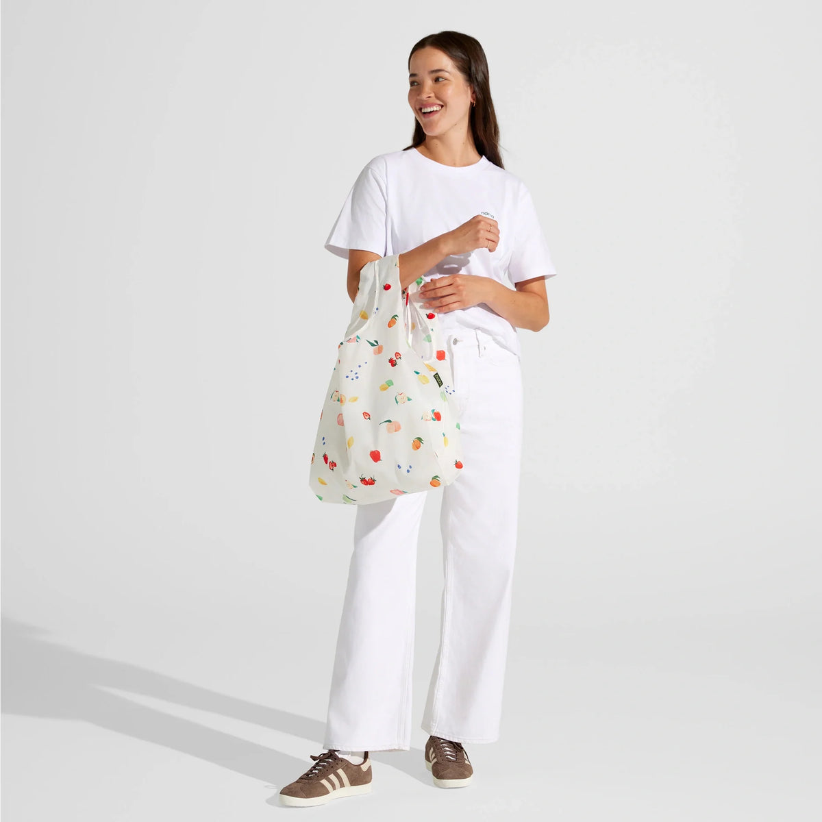 A woman in a white t-shirt and white pants stands holding the Nama + Baggu Standard Tote Bag, which is decorated with a colorful fruit pattern suggesting fresh produce inside. She is smiling and wearing brown sneakers.