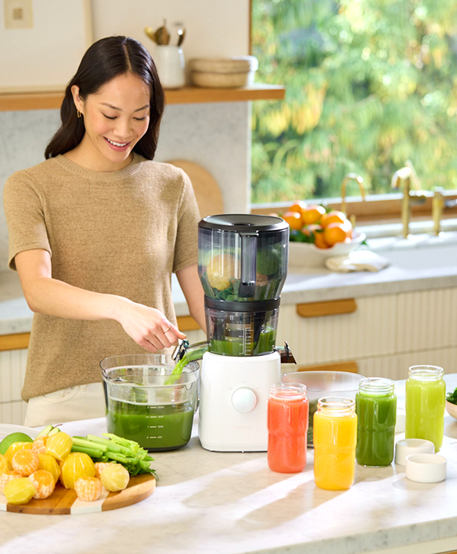 A woman in a beige top uses a juicer in a bright kitchen, pouring green juice into a pitcher. Colorful jars of juice and fresh citrus fruit are on the counter.