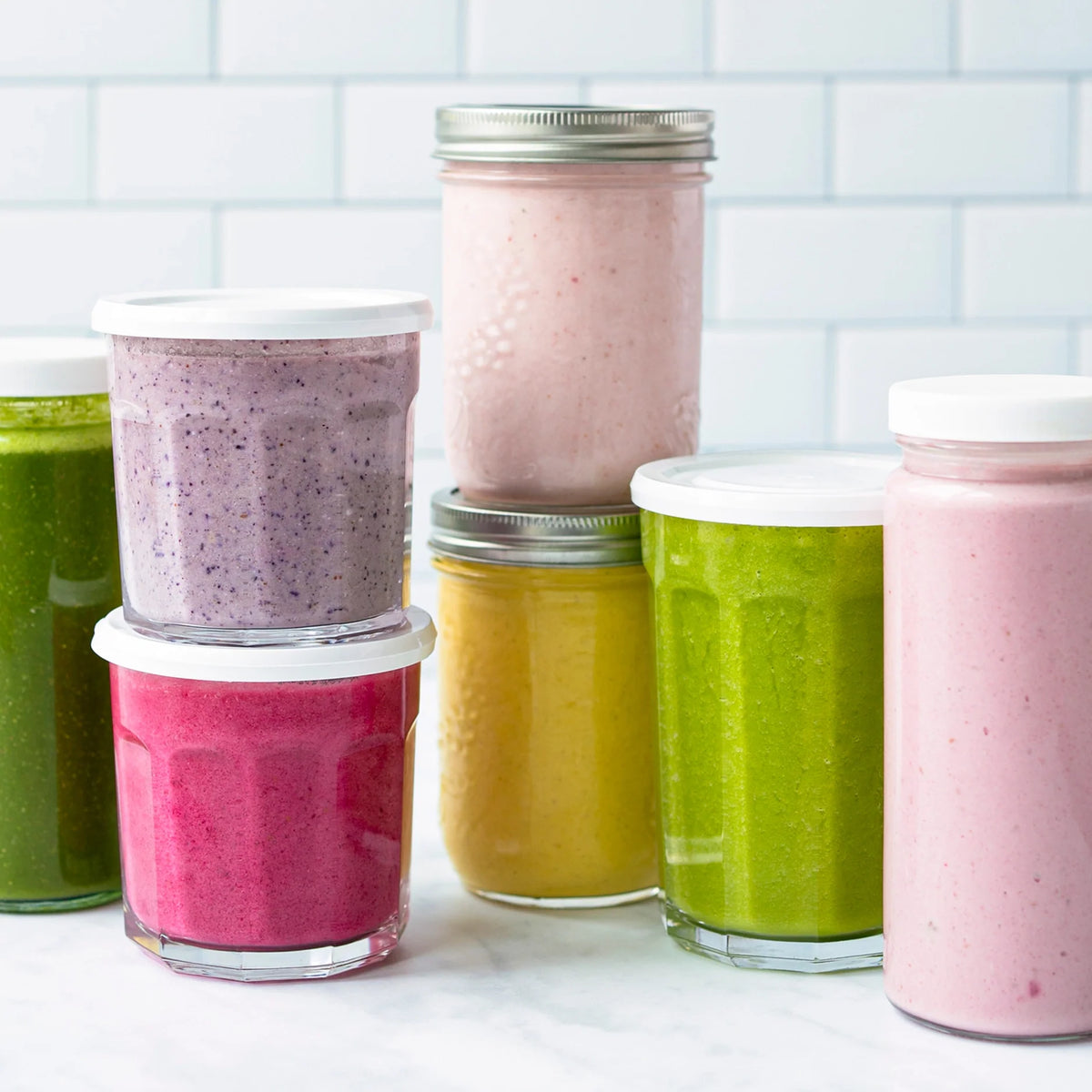 Assorted glass jars filled with colorful smoothies—pink, green, yellow, purple, and red—are arranged on a white countertop in front of a tiled white background.