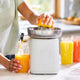 Using the Citrus Attachment, a person squeezes an orange, dispensing fresh juice into a glass. Two more filled glasses sit on the kitchen counter nearby, while sunlight streams through a window in the background.