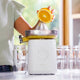 A person uses the Citrus Attachment to squeeze a halved orange, with juice flowing into a glass. Several empty glasses are lined up on a bright kitchen counter.