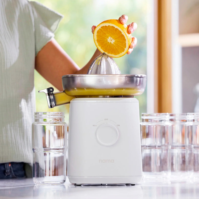 A person uses the Citrus Attachment to squeeze a halved orange, with juice flowing into a glass. Several empty glasses are lined up on a bright kitchen counter.