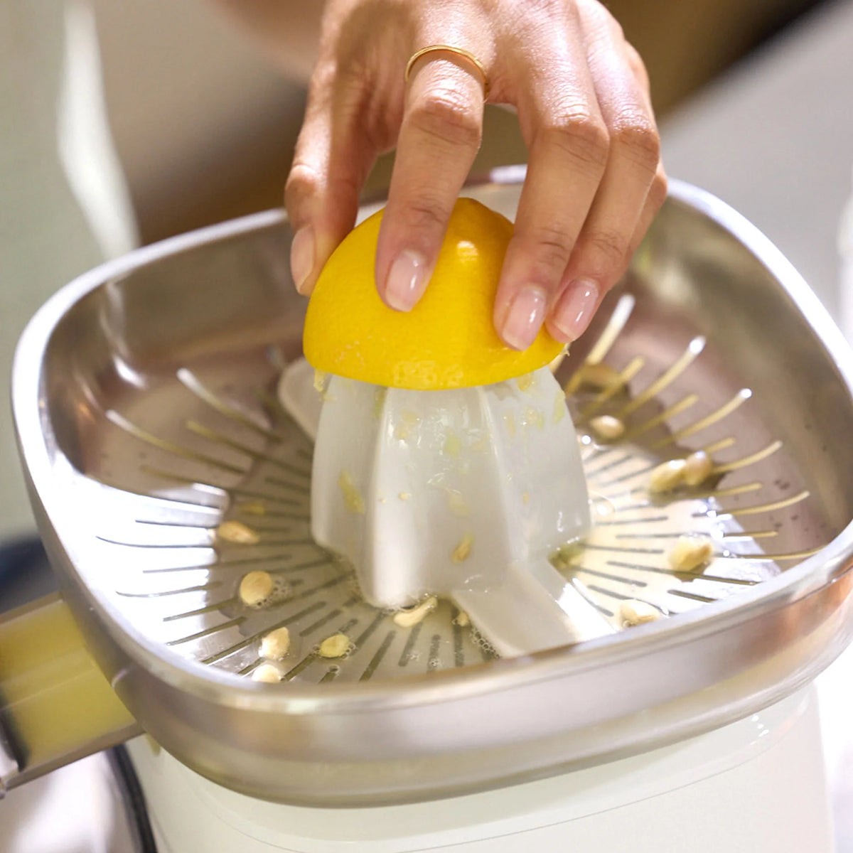 A hand presses half a lemon onto the Citrus Attachment, extracting juice from the fruit while lemon seeds and pulp collect on the metal strainer. The person wears a simple gold ring on their finger.