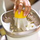 A hand presses half a lemon onto the Citrus Attachment, extracting juice from the fruit while lemon seeds and pulp collect on the metal strainer. The person wears a simple gold ring on their finger.