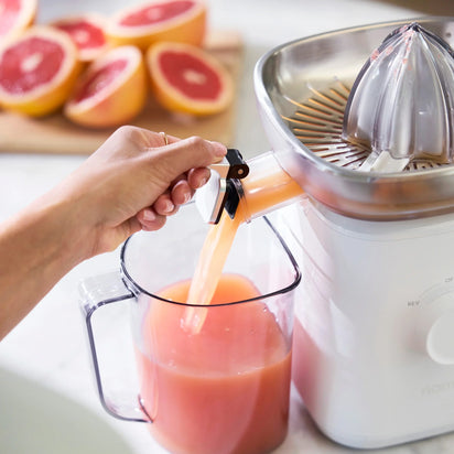 A hand pours fresh grapefruit juice from the Citrus Attachment into a clear pitcher. Sliced grapefruits are visible on a wooden cutting board in the background, highlighting how easy it is to juice citrus fruits with this Citrus Attachment.