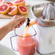 A hand pours fresh grapefruit juice from the Citrus Attachment into a clear pitcher. Sliced grapefruits are visible on a wooden cutting board in the background, highlighting how easy it is to juice citrus fruits with this Citrus Attachment.