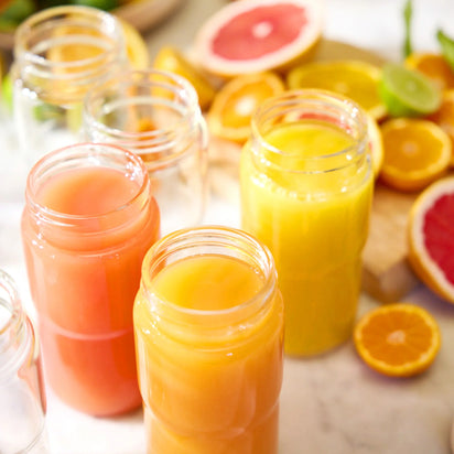 Three glass jars filled with orange and pink juice are on a table, surrounded by sliced oranges, grapefruits, and limes, creating a fresh and vibrant setting that highlights the delicious results you can achieve when you juice citrus fruits with the Citrus Attachment.