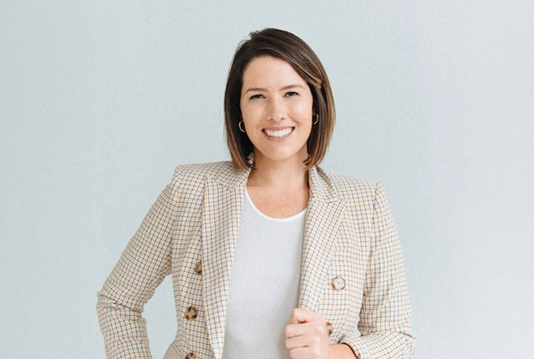A smiling woman with short brown hair stands against a light background, wearing a plaid blazer over a white top with one hand holding her blazer lapel.