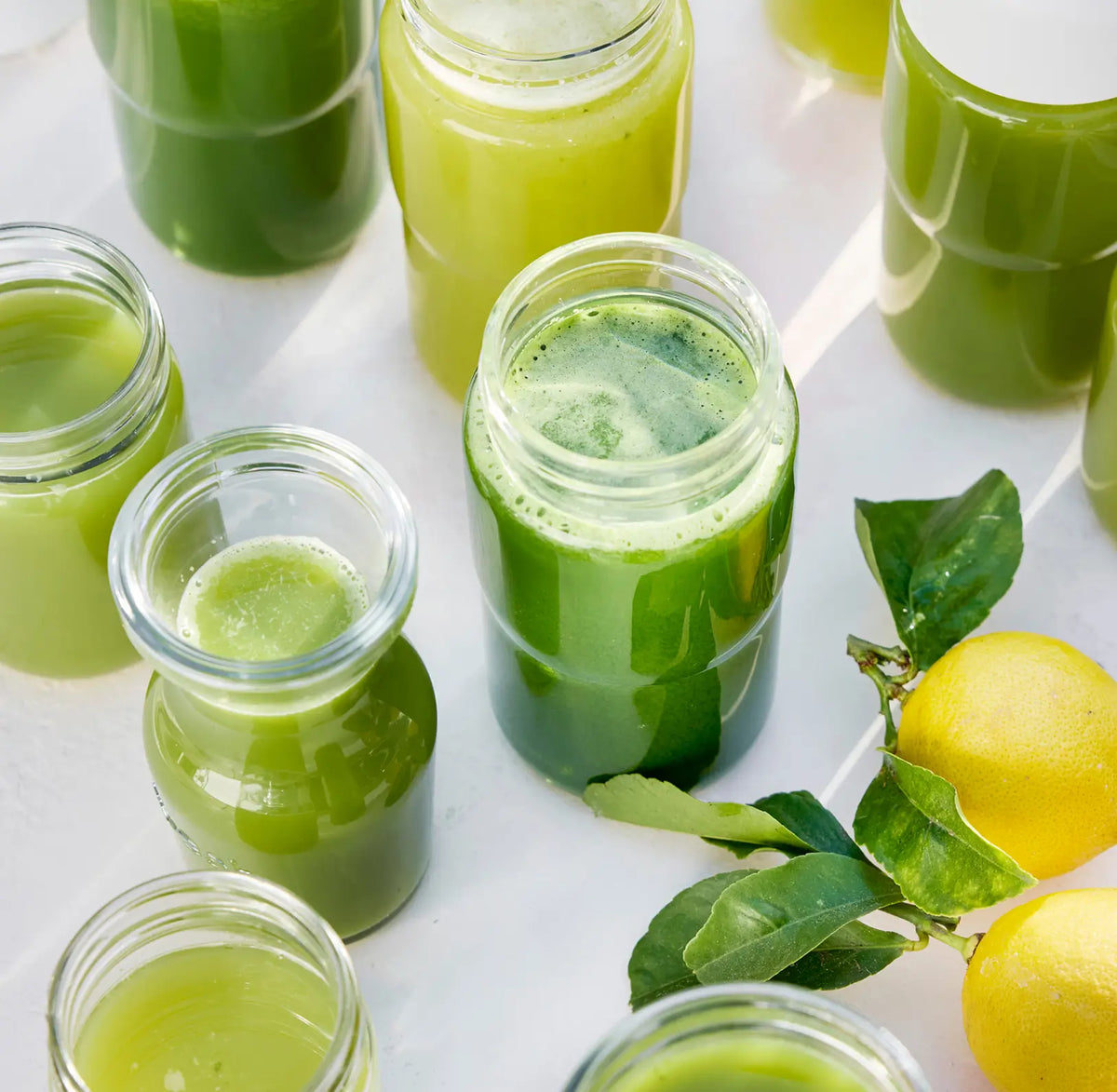 Several glass jars filled with green juice are arranged on a white surface next to two whole yellow lemons with green leaves still attached.
