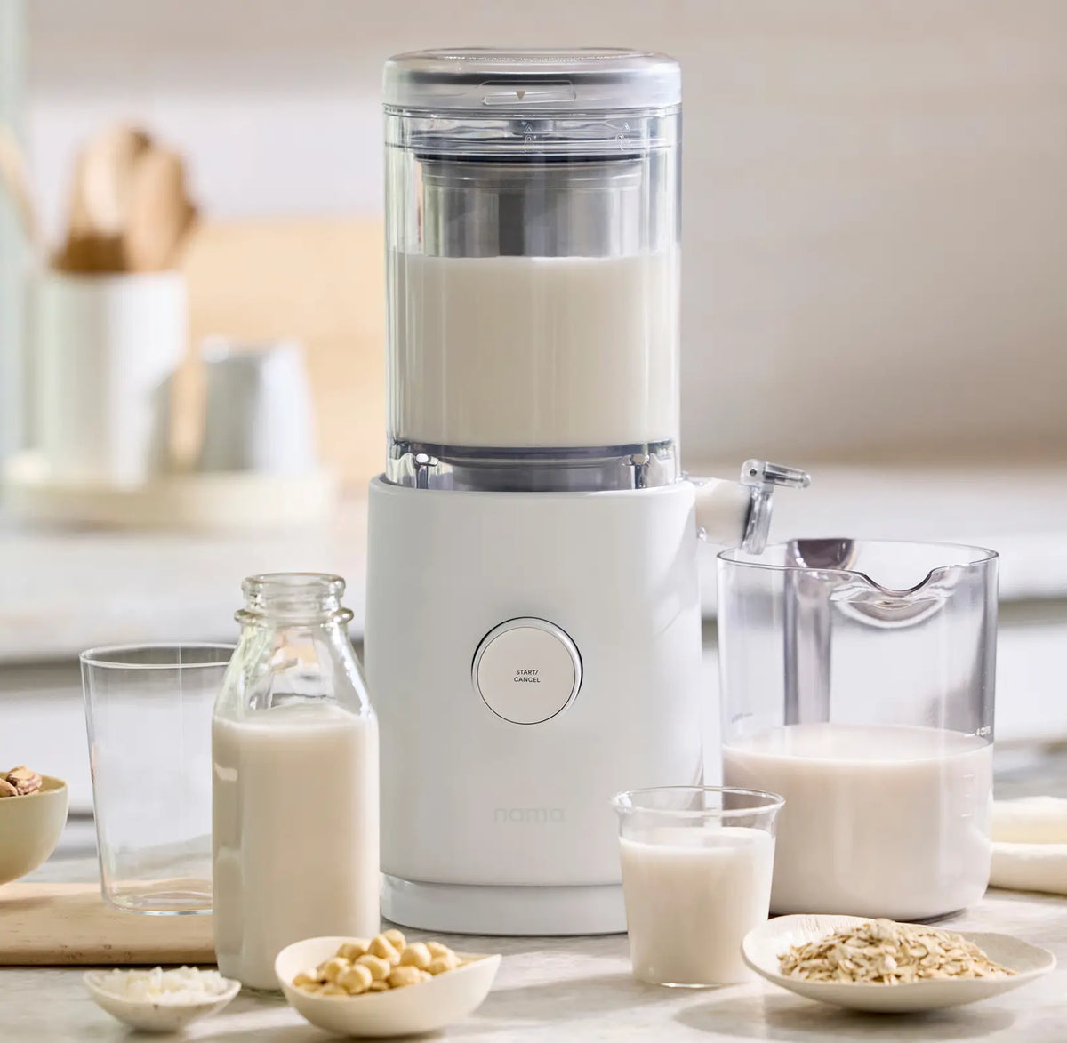 A blender filled with plant-based milk stands on a counter, surrounded by a glass bottle, a clear pitcher, a glass of milk, and bowls of oats and nuts. The background features a softly lit kitchen setting.