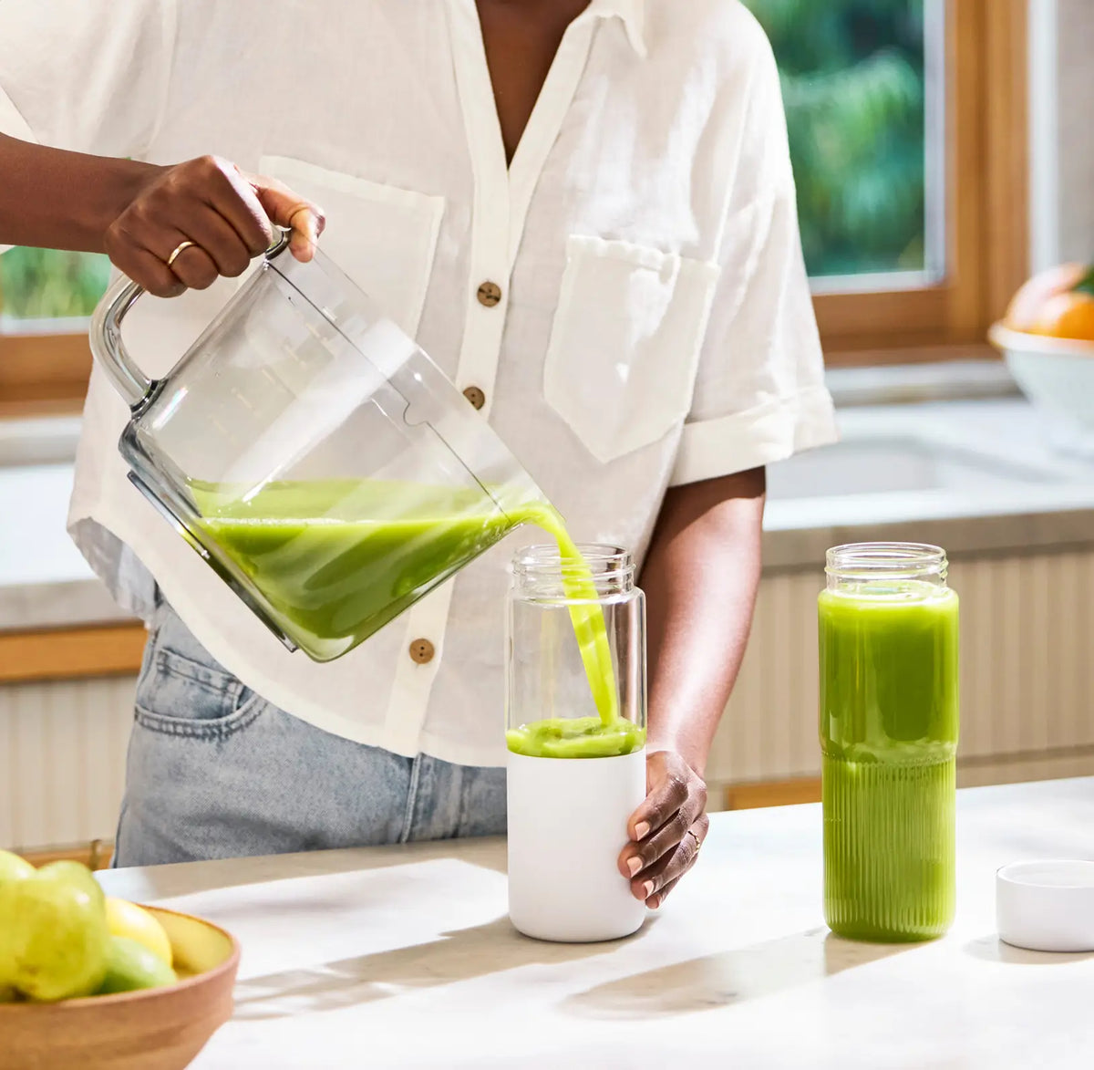 A person in a white shirt pours green juice from a blender into a travel bottle in a bright kitchen. Another filled bottle and a bowl of green apples are on the counter.