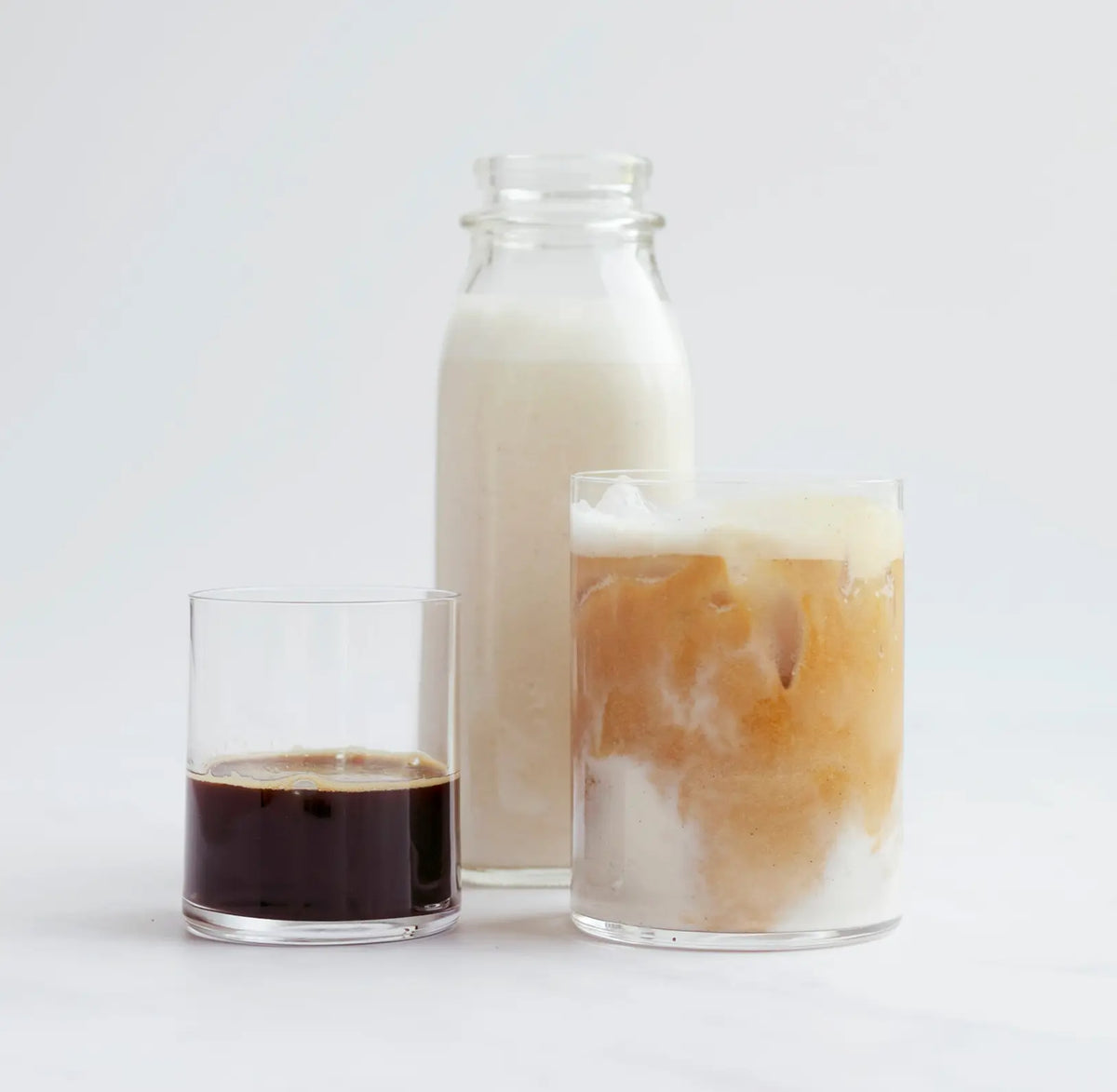 Three glasses: one with black coffee, one with iced coffee and milk, and one tall bottle filled with milk, all set against a plain white background.