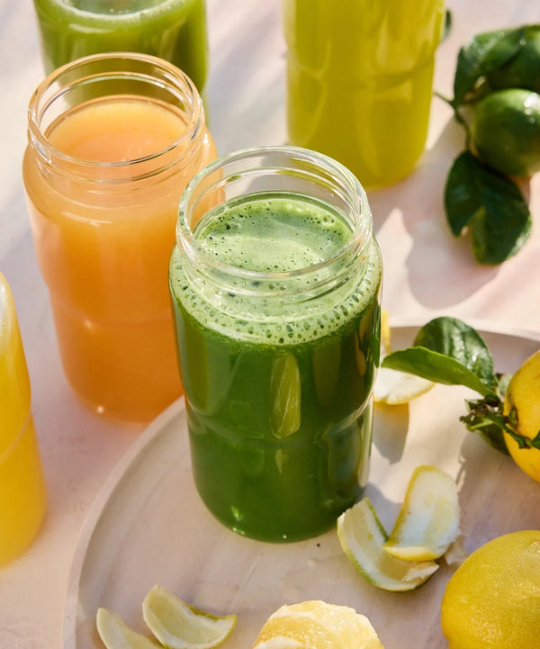 Three jars of colorful juices—green, orange, and yellow—sit on a light surface near lemons, limes, and citrus peels, with bright natural lighting highlighting the fresh ingredients.
