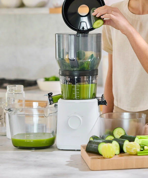A person adds a cucumber slice to a white juicer filled with green juice, with cut cucumbers and limes on a board nearby and a measuring cup collecting juice below.