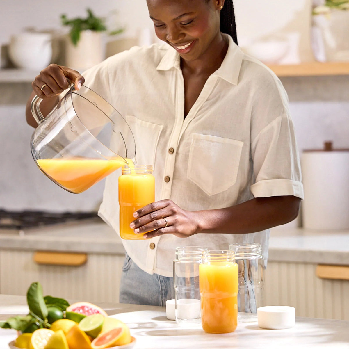 A woman smiles while pouring orange juice from a Glass Bottle Set into a jar with a mason jar lid in a bright kitchen. Sliced citrus fruits are on the counter nearby.