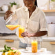 A woman smiles while pouring orange juice from a Glass Bottle Set into a jar with a mason jar lid in a bright kitchen. Sliced citrus fruits are on the counter nearby.