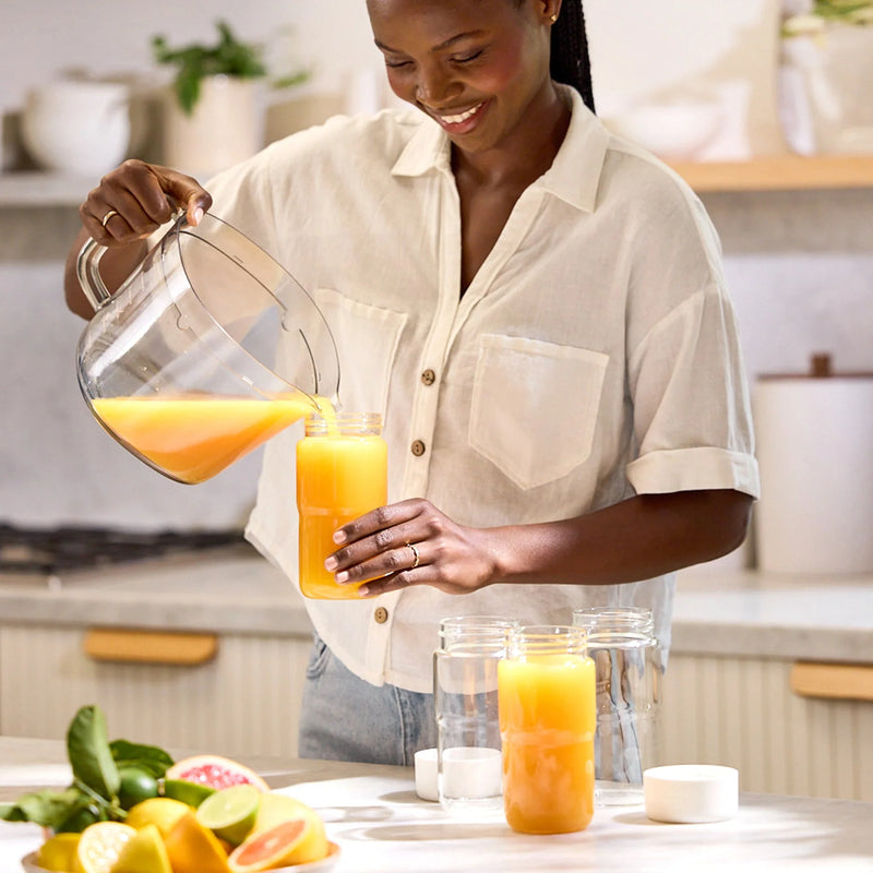 A woman smiles while pouring orange juice from a Glass Bottle Set into a jar with a mason jar lid in a bright kitchen. Sliced citrus fruits are on the counter nearby.