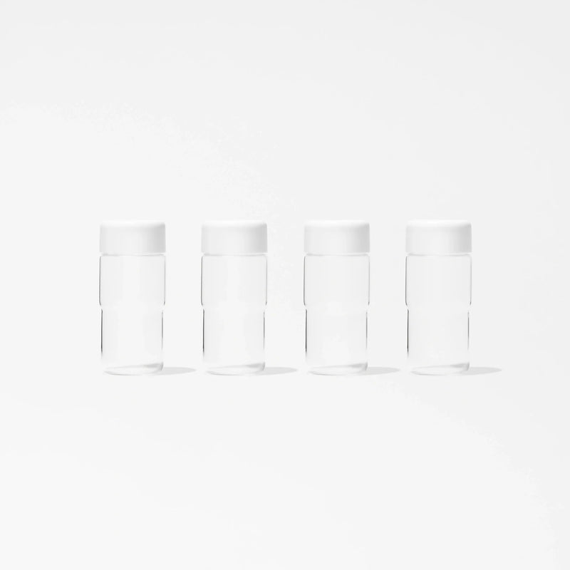 A set of four identical, clear cylindrical glass bottles from the Glass Bottle Set, each topped with a white mason jar lid, are displayed in a straight line on a plain white background. Evenly spaced and matching in design, these bottles are ideal for organized juice storage.