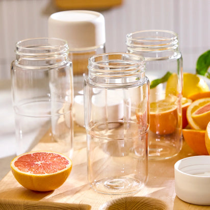 Three empty, clear bottles from the Glass Bottle Set with white mason jar lids sit on a wooden surface, surrounded by halved and sliced citrus fruits, including grapefruits and lemons. Bright natural light highlights the scene, inspiring fresh ideas for juice storage and presentation.