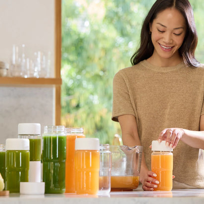 A woman smiles while sealing a bottle of orange juice with a mason jar lid; several filled bottles from the Glass Bottle Set, containing green and orange juice, are arranged on a kitchen counter beside her, ready for convenient juice storage.