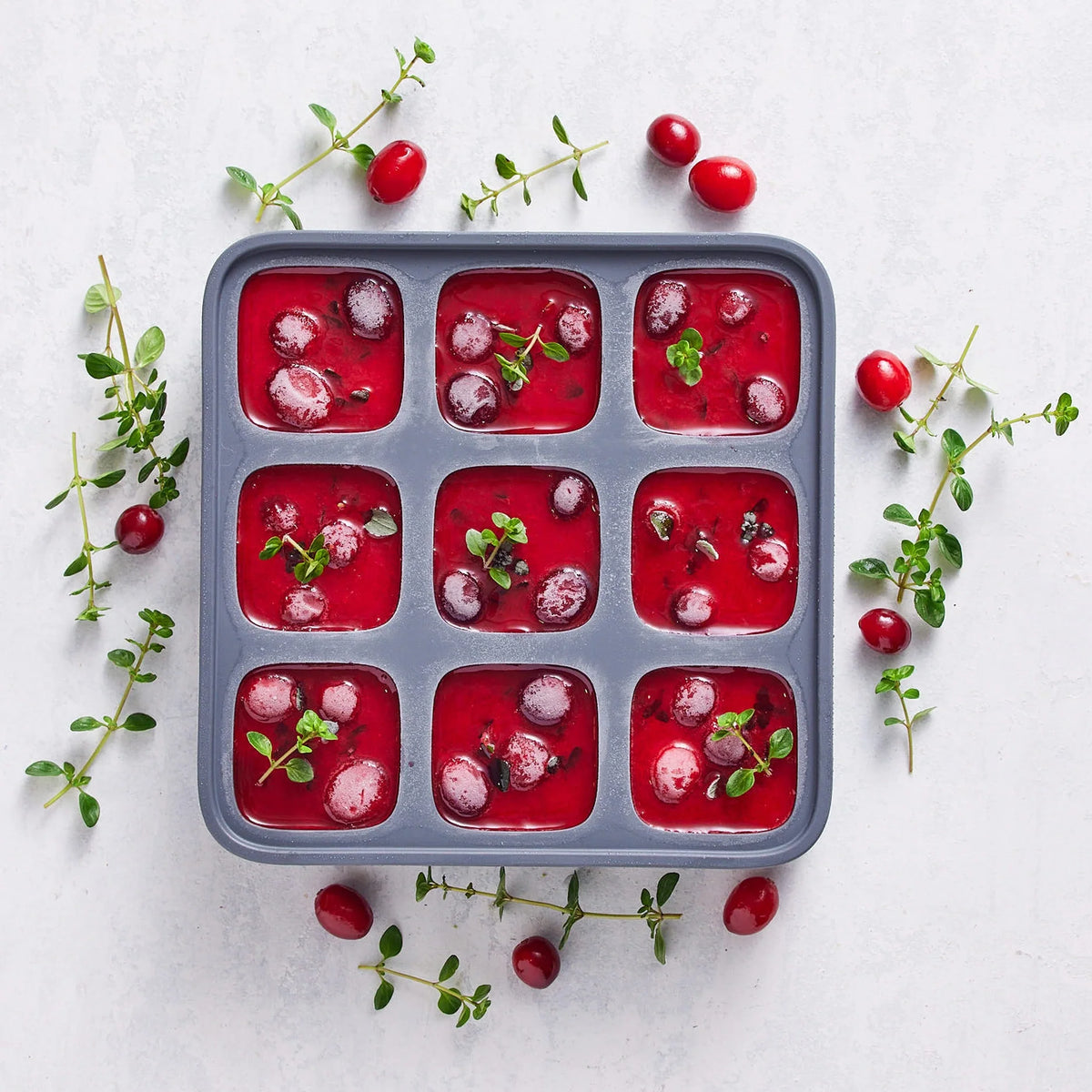 An ice cube tray filled with nine compartments of red cranberry sauce, each garnished with whole cranberries and green herbs, sits on a light surface scattered with additional cranberries and herbs.