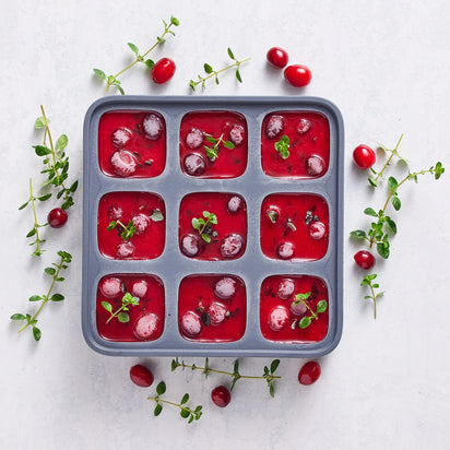 An ice cube tray filled with nine compartments of red cranberry sauce, each garnished with whole cranberries and green herbs, sits on a light surface scattered with additional cranberries and herbs.