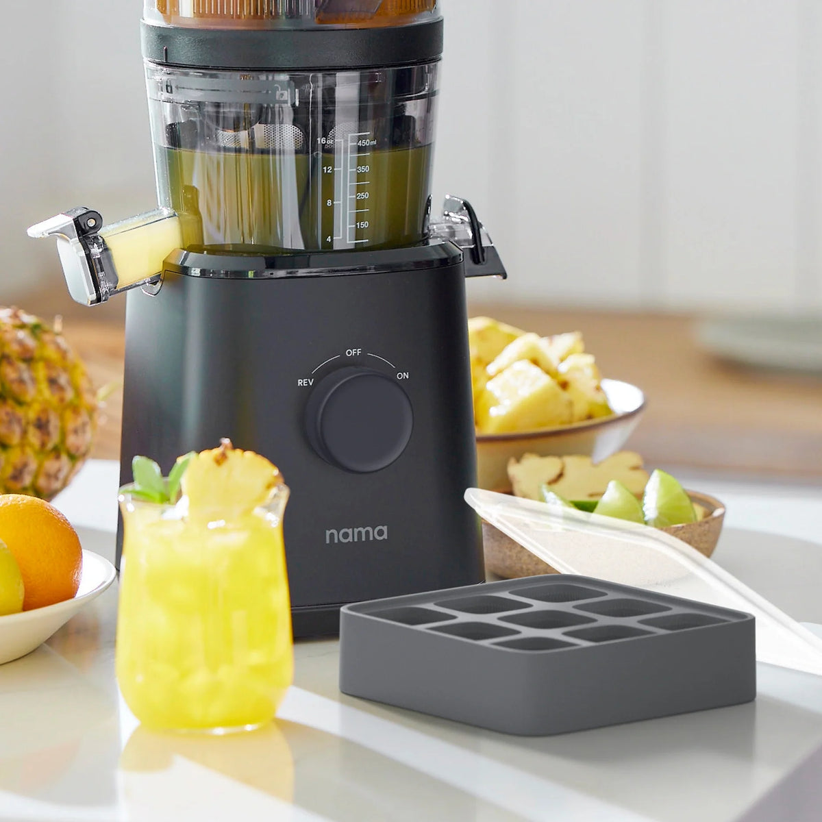 A black juicer filled with chopped pineapple sits on a counter next to a glass of yellow juice with ice, an Ice Cube Tray, pineapple chunks, and citrus fruits.