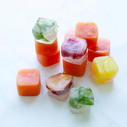 Ice cubes made with the Ice Cube Tray are stacked and scattered on a white surface, featuring vibrant colors from fruit and herbs. Some cubes display visible mint leaves, while others appear orange, yellow, or red.
