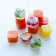 Ice cubes made with the Ice Cube Tray are stacked and scattered on a white surface, featuring vibrant colors from fruit and herbs. Some cubes display visible mint leaves, while others appear orange, yellow, or red.
