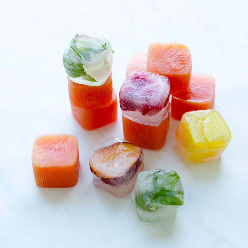 Ice cubes made with the Ice Cube Tray are stacked and scattered on a white surface, featuring vibrant colors from fruit and herbs. Some cubes display visible mint leaves, while others appear orange, yellow, or red.