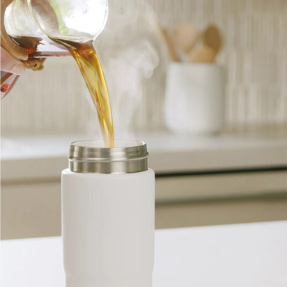 A hand pours steaming hot coffee from a glass container into an Insulated On-the-Go Bottle with a ceramic lining on a kitchen counter, while blurred utensils sit in the background.