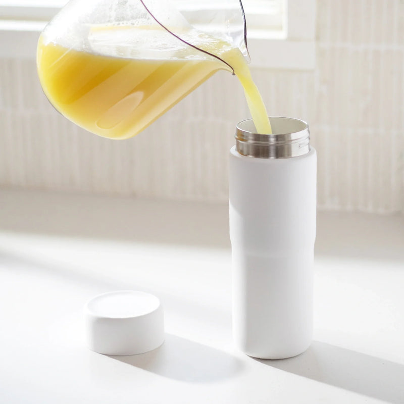 A glass pitcher pours yellow juice into the Insulated On-the-Go Bottle, which features a ceramic lining and is set on a white surface with its cap placed nearby. Soft light streams through a window in the background.