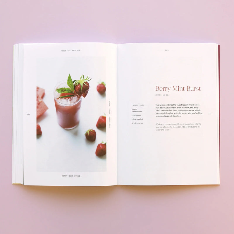 An open Juice the Rainbow Book displays a photo of a pink smoothie garnished with mint and strawberries, with fresh strawberries scattered around. The opposite page features the Berry Mint Burst recipe on a light pink background, showcasing the vibrant flavors and plant-based nutrition found in creative juice recipes.