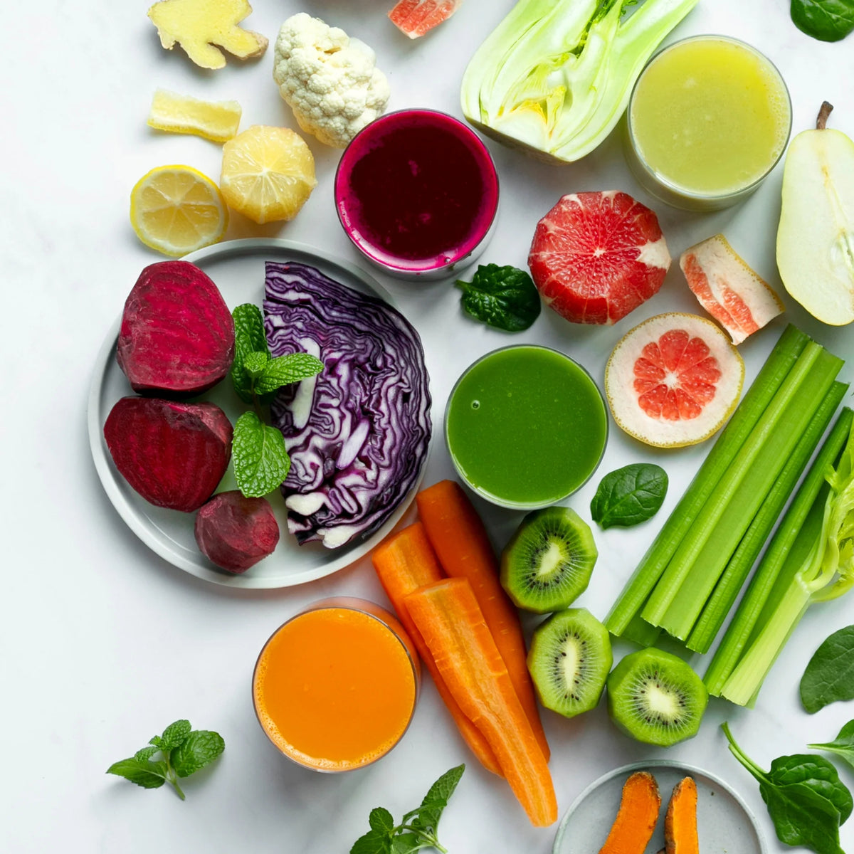 Four glasses of fresh juice from the “Juicing for Balanced Blood Sugar” collection are arranged on a white surface, surrounded by colorful fruits and vegetables such as beets, kiwi, carrots, grapefruit, celery, ginger, cabbage, lemon, and greens.