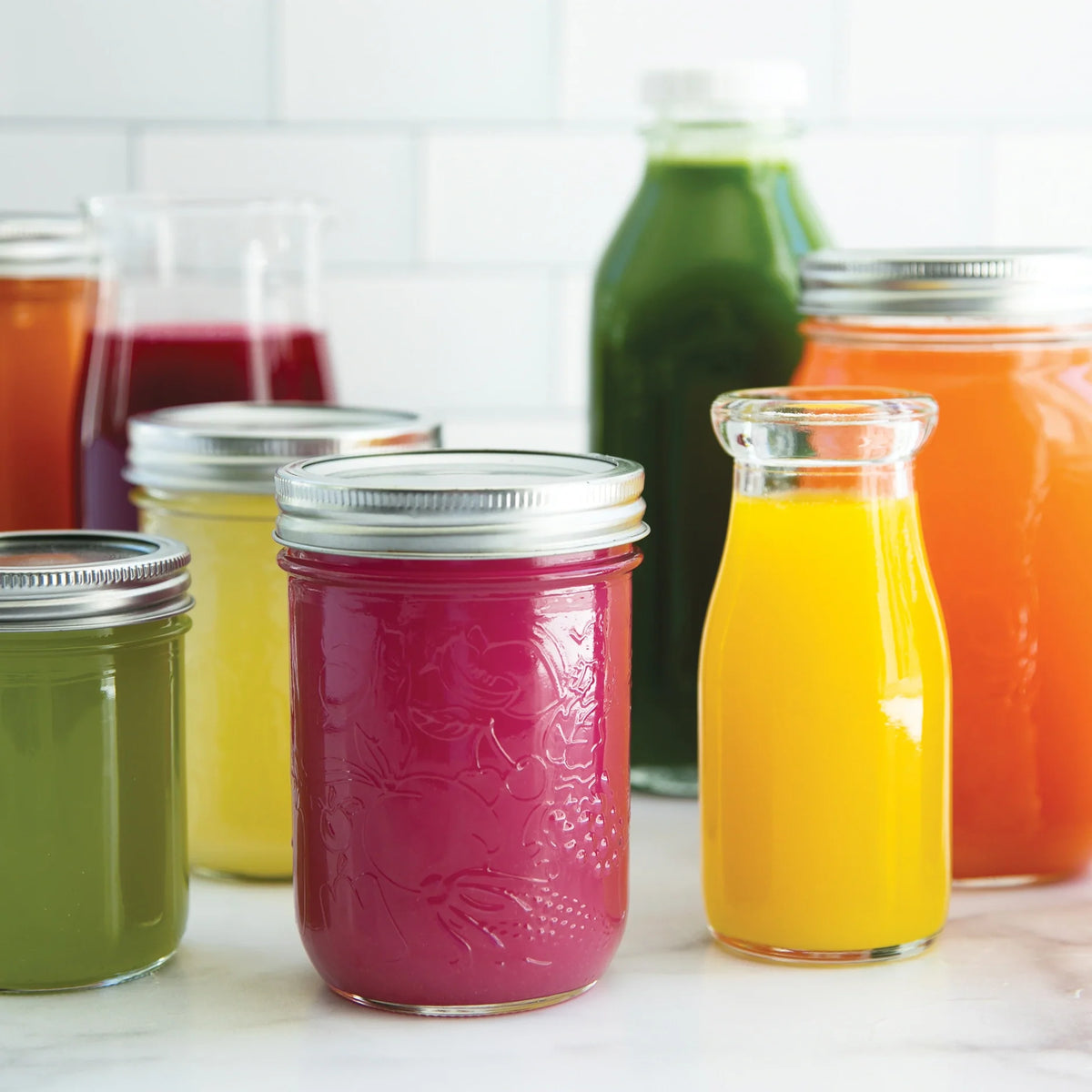 Glass jars and bottles brimming with vibrant orange, yellow, green, red, and purple juices—some sealed with metal lids, others open—are arranged on a white surface before a backdrop of white tiles. Featuring recipes from Juicing for Beginners, these colorful drinks are the perfect addition to plant-based meal plans or anyone seeking inspiration from a comprehensive juicing guide to add freshness to their daily routine.