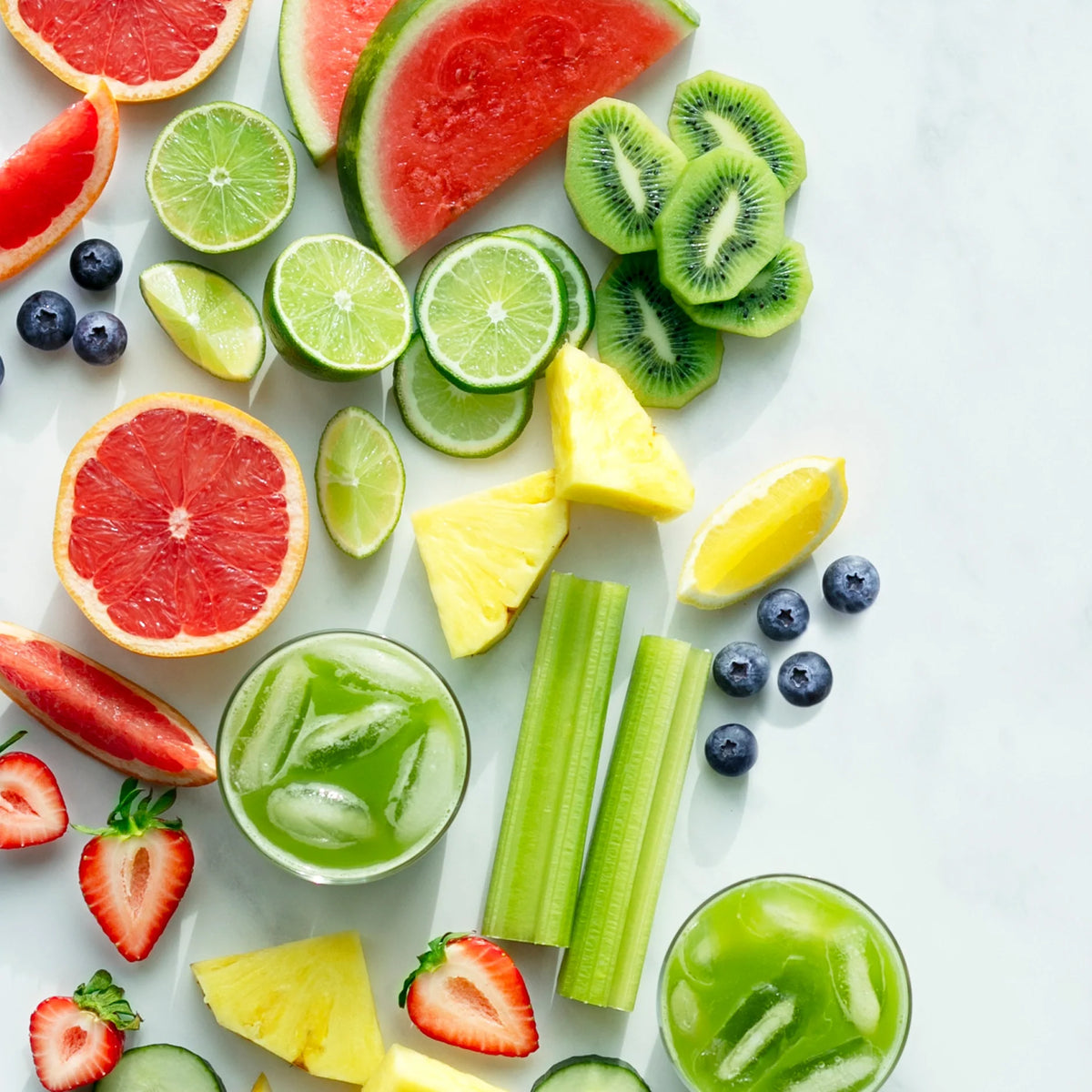 An assortment of fresh fruit—including watermelon, kiwi, lime, grapefruit, pineapple, strawberries, and blueberries—plus celery is artfully arranged with two glasses of Skin Health green juice on a light background.